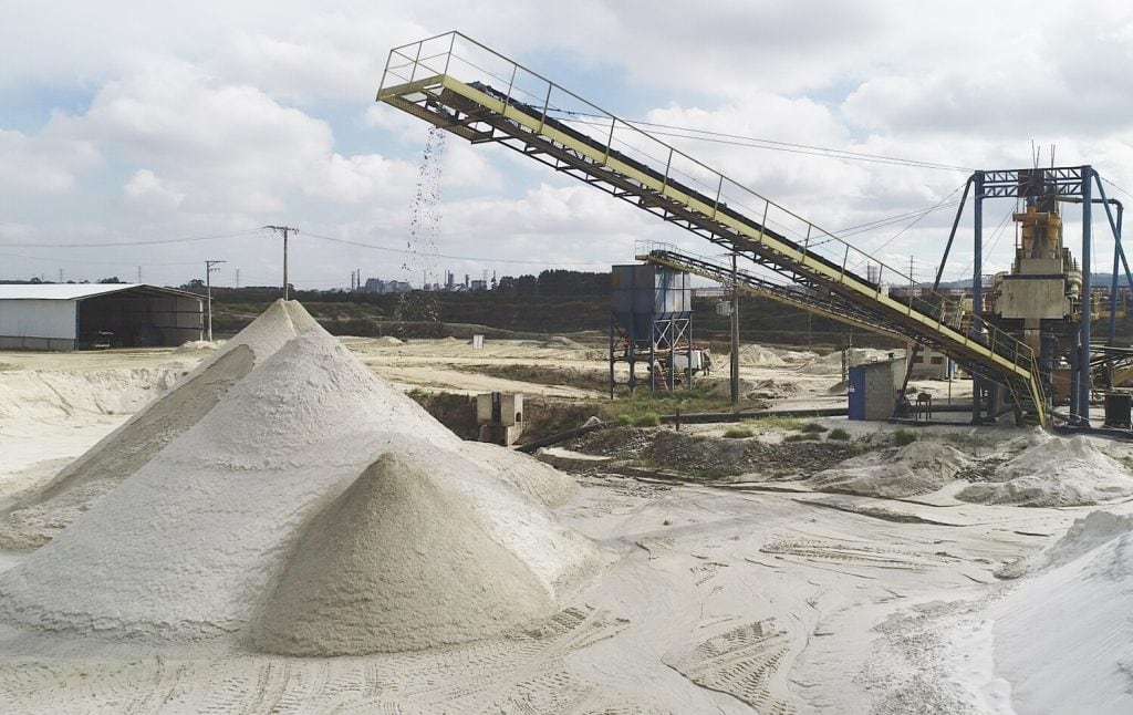 Areia caindo da esteira transportadora sobre uma pilha cônica, com a estrutura da usina ao fundo